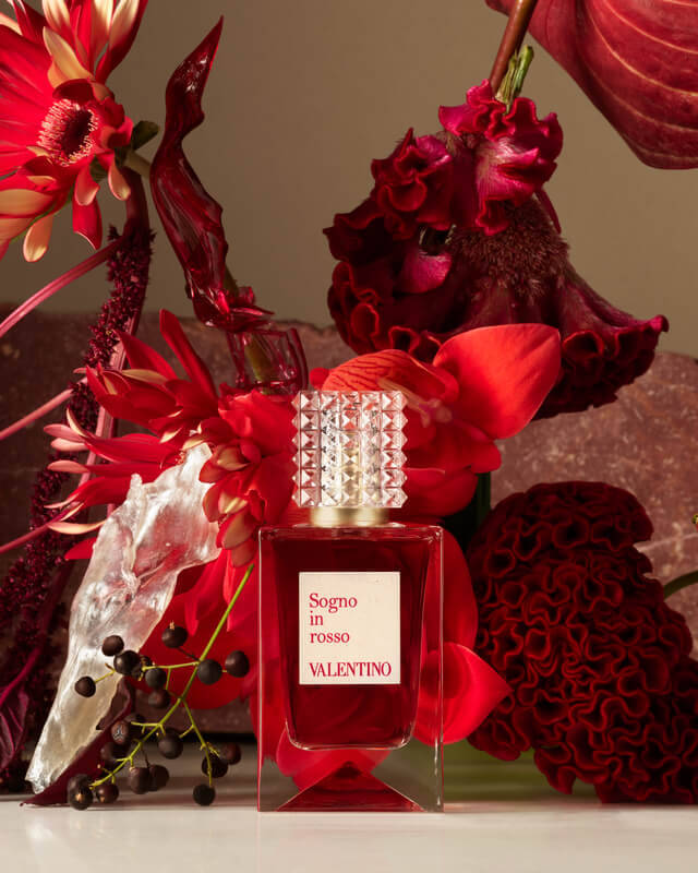 Een geurflacon van Valentino omringd door verschillende rode bloemen en planten, waaronder rozen en anthuriums, op een lichte achtergrond.