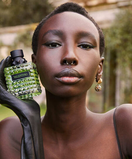 Model met kort haar en zwarte handschoen houdt een fles Valentino-parfum vast in een groene omgeving.