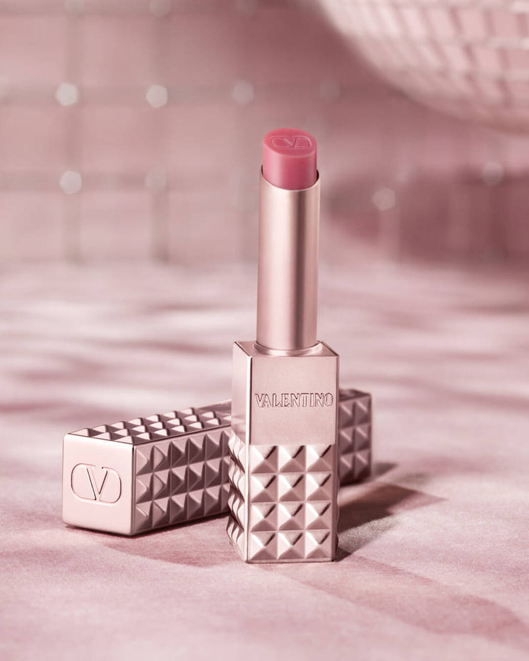 Een rosegoudkleurig Valentino Spike lipproduct met een roze punt staat rechtop, met de bijpassende bezaaide huls ernaast. Beide items liggen op een zachte, glinsterende roze achtergrond die een gevoel van glamour oproept.