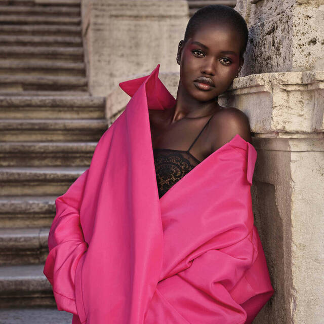Een model met een stralende huid draagt een fuchsia oversized jas en zwarte lingerie, staande op een stenen trap.